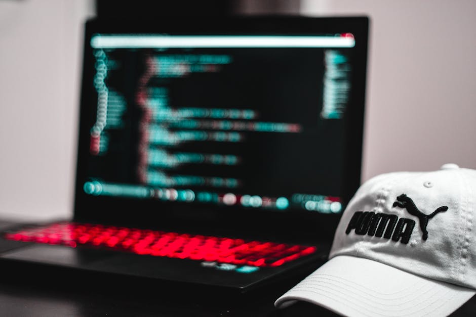 The image shows a person working at a desk with two computer monitors in an office environment. The individual, whose back is visible, is wearing glasses and has short hair. The primary monitor on the