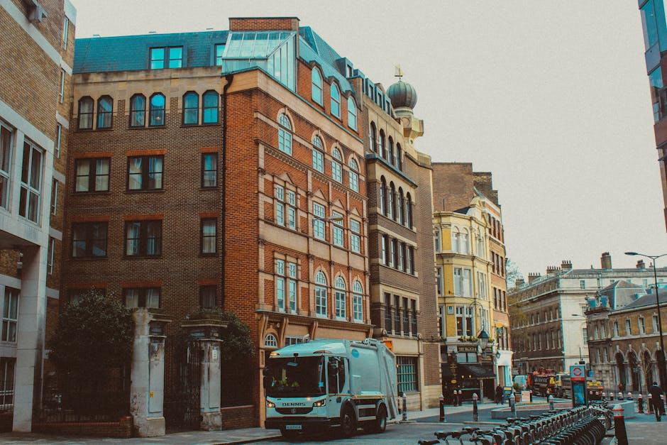 A city street scene featuring a collection of multi-storey buildings with distinctive architectural details, including large arched windows and decorative brickwork. The buildings display a mix of bri